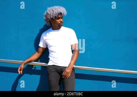 Black man with afro hair dressed in white leaning against a blue wall. Afro hair concept. Black man in white t-shirt. Stock Photo