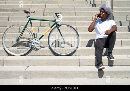 Black man talking on the phone next to his bike. man sitting on the stairs and calling with his cell phone. Stock Photo