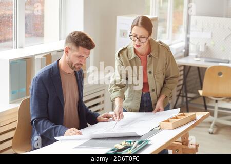 Waist up portrait of two architects pointing at blueprints and discussing work while standing by drawing desk in office lit by sunlight , copy space Stock Photo