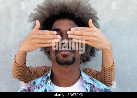 Anonymous friend covering eyes of stylish ethnic male with Afro hairstyle near concrete wall Stock Photo