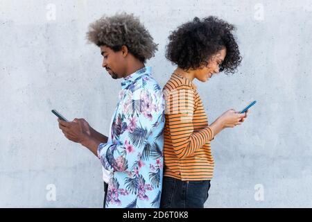 Side view of content multiracial couple of partners in trendy clothes with Afro hairstyle standing near wall with cellphones Stock Photo