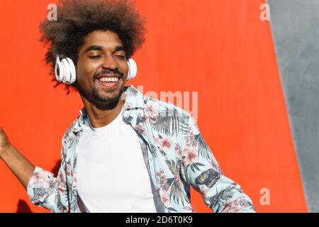 Young masculine bearded African American male with Afro hairstyle and closed eyes listening to music in headphones near red wall in town Stock Photo