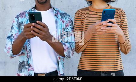 Cropped unrecognizable multiracial couple of partners in trendy clothes with Afro hairstyle standing near wall with cellphones Stock Photo