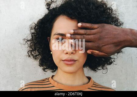 Anonymous friend covering eye of crop stylish ethnic female with curly hair hairstyle looking at camera near concrete wall Stock Photo