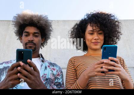 Content multiracial couple of partners in trendy clothes with Afro hairstyle standing near wall with cellphones Stock Photo