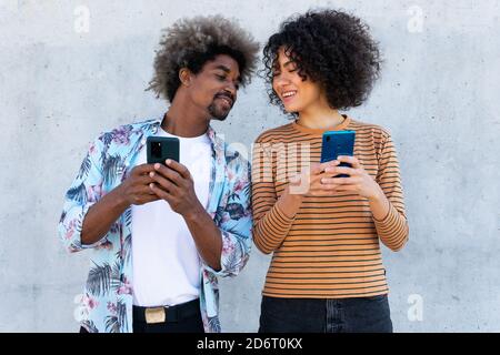 Content multiracial couple of partners in trendy clothes with Afro hairstyle standing near wall with cellphones Stock Photo