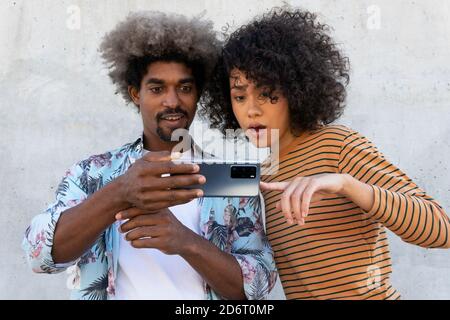 Smiling stylish black man near surprised ethnic female partner with Afro hairstyle pointing with finger at cellphone on street Stock Photo