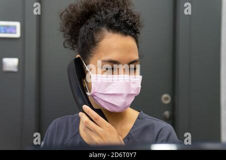 Adult ethnic female medic in mask and uniform answering phone call while working in modern clinic Stock Photo
