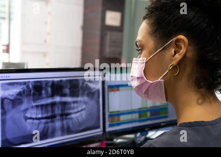 Side view of black female doctor in medical mask examining X ray on monitor of computer while working in modern dental clinic Stock Photo