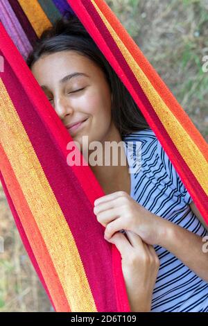 Female lying and swinging in hammock in green garden on sunny day while having sun and closed eyes Stock Photo