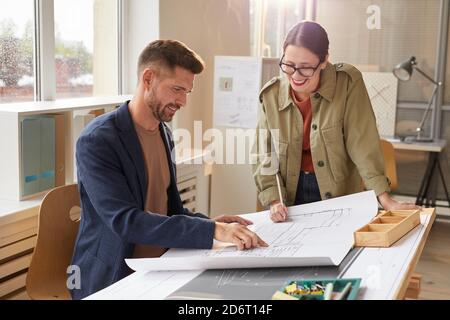 Waist up portrait of two smiling architects pointing at blueprints and discussing work while standing by drawing desk in office lit by sunlight , copy space Stock Photo