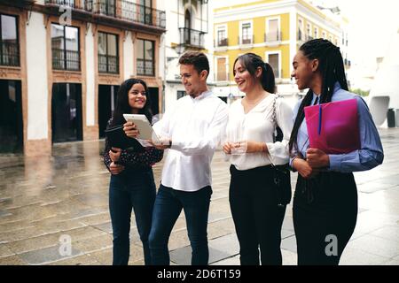Young smiling man sharing tablet with stylish multiracial partners on city street in daylight Stock Photo