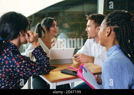 Cheerful multiethnic colleagues talking while looking at each other and sitting at table with netbook in building Stock Photo