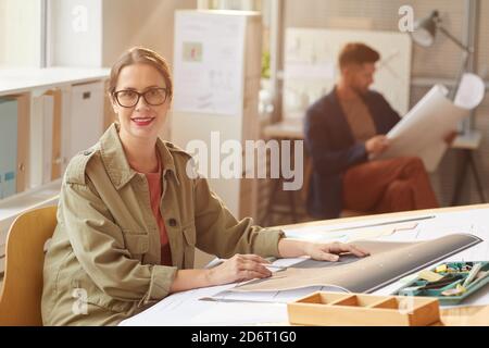 Portrait of young female architect looking camera and smiling while sitting at drawing desk in sunlight, copy space Stock Photo