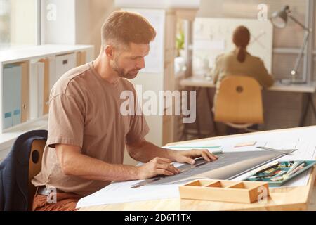 Side view portrait of mature bearded architect looking at blueprints while sitting at drawing desk in sunlight, copy space Stock Photo