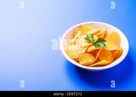 Fried corrugated golden potato chips with parsley leaf in wooden bowl on blue background, angle view copy space Stock Photo