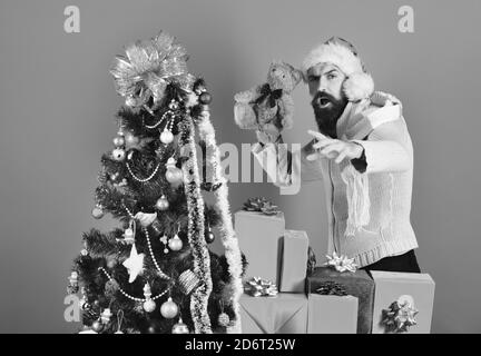 Man with beard ready to throw toy on blue background. Christmas gifts and surprise concept. Guy in red hat with xmas gift boxes and teddy bear in hands. Santa with red presents near decorated fir tree Stock Photo