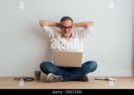 Full body delighted pleased mature male wearing casual clothes sitting on floor with contemporary laptop on laps Stock Photo