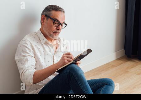 Focused mature male in casual wear and eyeglasses sitting on parquet at home and writing down thoughts in diary Stock Photo