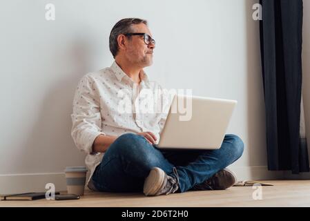 Contemplative senior male sitting with legs crossed on parquet with netbook on laps and cup of takeaway coffee in hand while looking away in thoughts Stock Photo