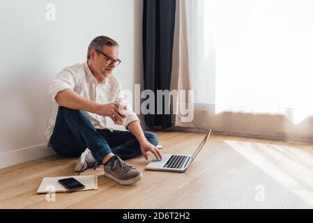 Contemplative senior male sitting with legs crossed on parquet with netbook on laps and cup of takeaway coffee in hand while looking away in thoughts Stock Photo