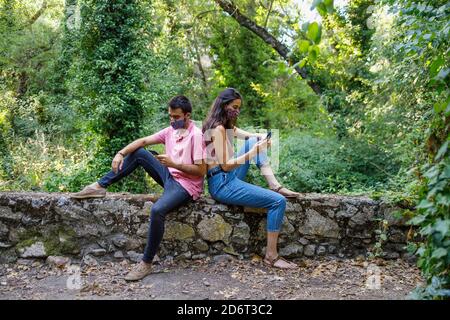 Side view of unrecognizable young Woman and man in face masks sitting back to back in green park and browsing smartphones during coronavirus Stock Photo