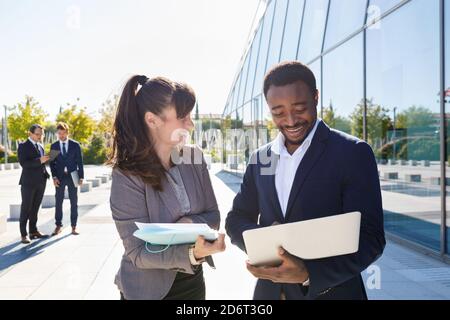 Focused young female manager in suit showing documents to African American male colleague standing near contemporary building with laptop on sunny day Stock Photo