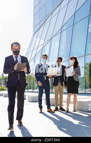 Full body of anonymous male manager in formal suit and medical mask using tablet while walking on street near diverse coworkers working on laptop Stock Photo