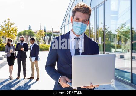 Unrecognizable young male manager in stylish suite and protective mask working on laptop while standing on street near building and diverse colleagues Stock Photo
