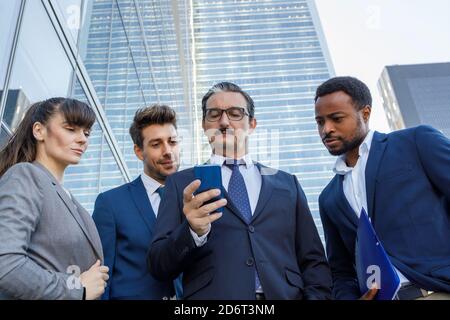 Group of happy multiracial colleagues in classic suites laughing while reading good news on smartphone standing together on street Stock Photo