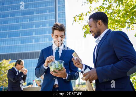 Positive young diverse coworkers in formal clothes smiling while having lunch break together on street on sunny day Stock Photo