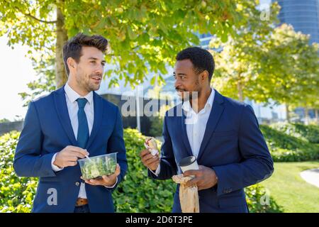 Positive young diverse coworkers in formal clothes smiling while having lunch break together on street on sunny day Stock Photo