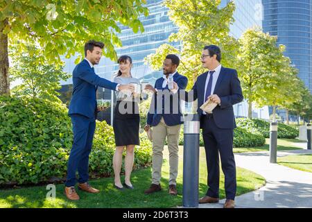 Cheerful multiracial coworkers in formal clothes standing in city park and clinking with coffee cups while celebrating successful agreement Stock Photo
