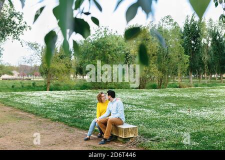 Tender girlfriend and boyfriend sitting on bench and cuddling while looking at each other and enjoying weekend together Stock Photo