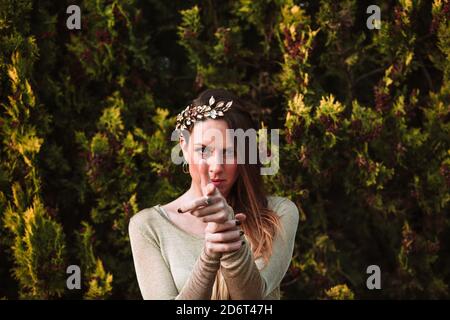 Confident young female wearing trendy hair wreath standing against abundant greenery while pointing finger gun and looking at camera Stock Photo