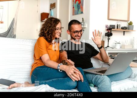 Cheerful young diverse couple in casual clothes and eyeglasses holding hands while sitting together on sofa and having video call on laptop Stock Photo