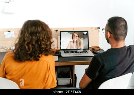 Cheerful young diverse couple in casual clothes and eyeglasses while sitting together and having video call on laptop Stock Photo