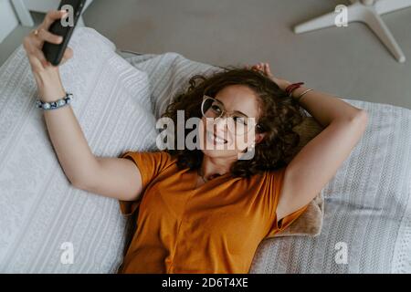 Top view of young barefooted lady with curly ginger hair in casual outfit lying on comfortable sofa and taking selfie on smartphone at home Stock Photo