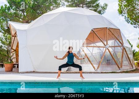 Smiling barefoot slim lady in sportswear standing on tiptoes with zen gesture near swimming pool while looking at camera Stock Photo