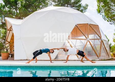Slim partners in sportswear standing in Camatkarasana pose while practicing yoga near swimming pool Stock Photo