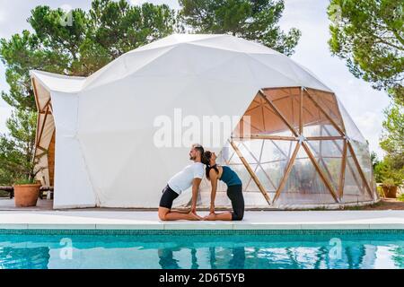 Side view of unrecognizable fit man and Woman standing in Ustrasana pose while practicing yoga and reflecting in swimming pool near tent Stock Photo