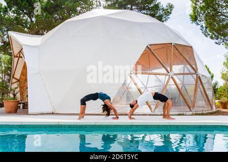 Slim partners in sportswear standing in bridge na pose while practicing yoga near swimming pool Stock Photo