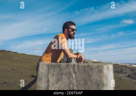 Side view of relaxed young ethnic guy in casual outfit and sunglasses leaning on wooden fence and admiring nature against blue sky Stock Photo