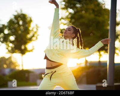Trendy young ethnic female dancer with stylish hairstyle in activewear performing freestyle movements in park at sunset Stock Photo