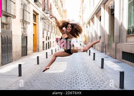 Full body of young fit ethnic female dancer with long curly hair doing grand jete position and smiling on paved street looking at camera Stock Photo