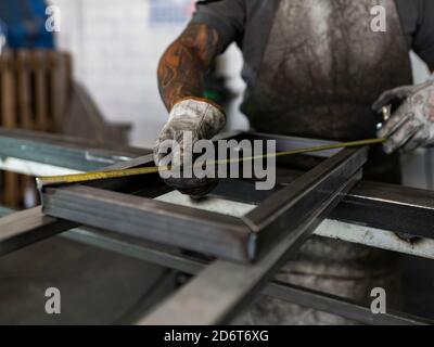 Unrecognizable male welder in dirty gloves and apron measuring frame with yellow metal tape and working at workbench in garage Stock Photo