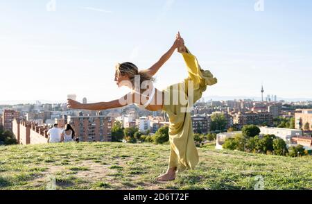 Side view full body barefoot female in bra and loose trousers performing Dancers Pose and outstretching hand with cup of coffee while practicing yoga Stock Photo