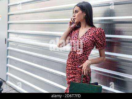 Young charming female standing near metal wall in city speaking on cellphone and communicating with friend on social media Stock Photo