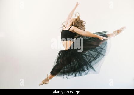 Side view of ballerina in black tutu jumping in splits with outstretched arms on white background in studio Stock Photo