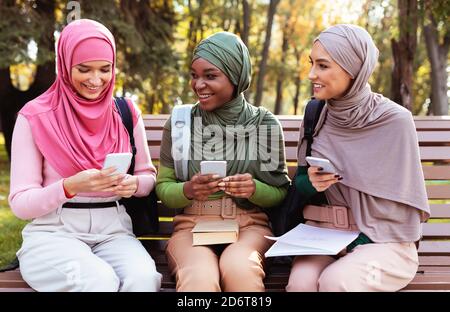 Three Muslim Students Women Using Smartphones Sitting On Bench Outdoors Stock Photo
